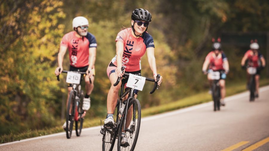 A group of cyclists riding on a paved road surrounded by trees, with a woman in the foreground wearing a helmet and sunglasses, smiling while cycling. Other riders follow behind, all wearing matching jerseys with race numbers.