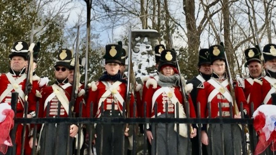 Group of historical reenactors wearing red military uniforms with black hats and carrying rifles, standing outdoors near a monument during a heritage or commemorative event.