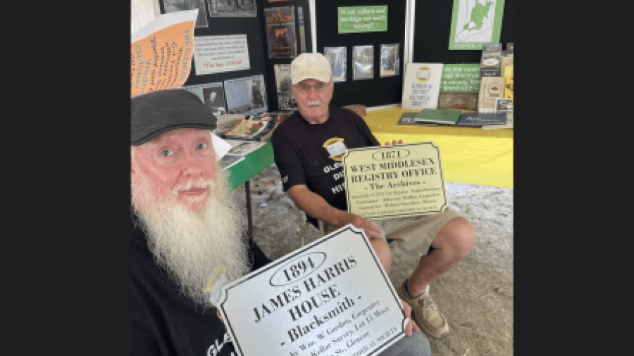 Photo of two men at a community heritage booth displaying historical materials, holding interpretive plaques for the 1894 James Harris House and the 1871 West Middlesex Registry Office, with informational displays about local history in the background.