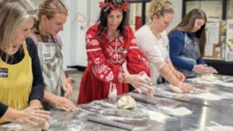 A group of participants working together at a cooking workshop, kneading dough and preparing pierogies on a flour-covered table, guided by an instructor wearing a traditional red Ukrainian-style dress and floral head wreath.
