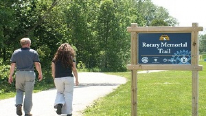 rotary trail with two passengers walking on a side walk/trail with the rotary memorial park sign beside them 