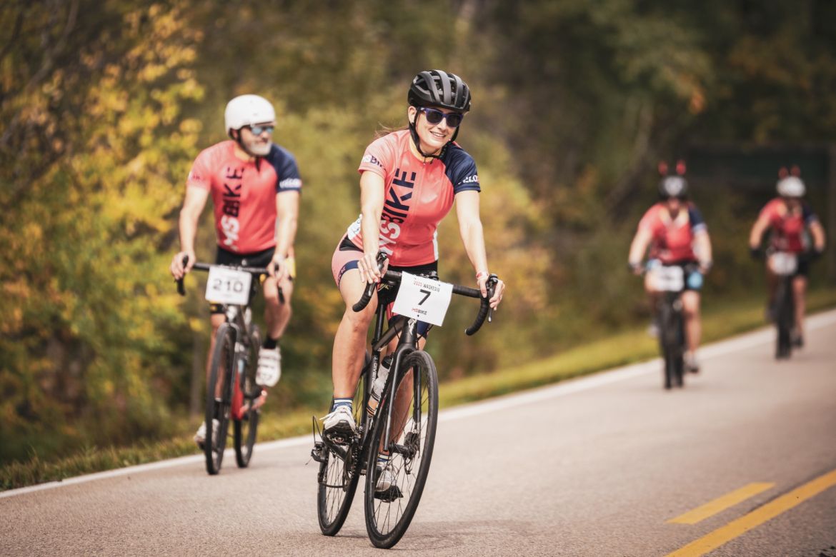 A group of cyclists riding on a paved road surrounded by trees, with a woman in the foreground wearing a helmet and sunglasses, smiling while cycling. Other riders follow behind, all wearing matching jerseys with race numbers.