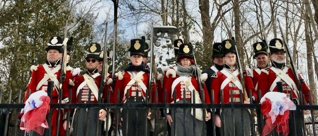 Group of historical reenactors wearing red military uniforms with black hats and carrying rifles, standing outdoors near a monument during a heritage or commemorative event.