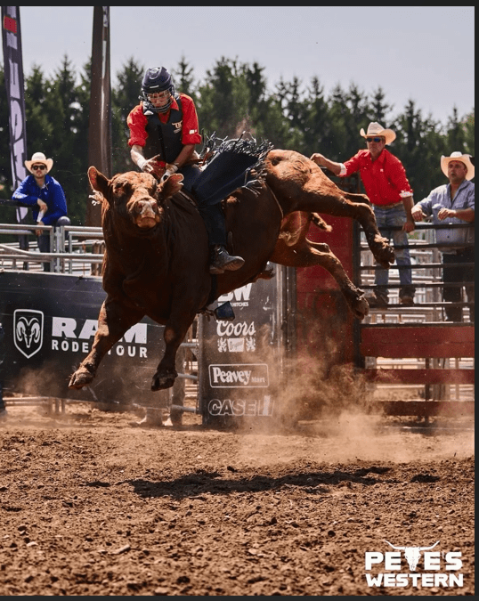 A rodeo rider wearing a helmet and protective vest competes in a bull riding event inside a dirt arena, gripping a bucking bull mid-jump while rodeo staff and spectators watch from behind the fence.
