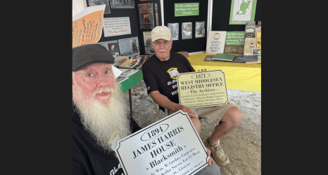 Photo of two men at a community heritage booth displaying historical materials, holding interpretive plaques for the 1894 James Harris House and the 1871 West Middlesex Registry Office, with informational displays about local history in the background.